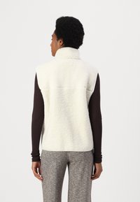 Vit mjuk fleeceväst med hög krage och utan ärmar, i kombination med en brun långärmad skjorta och grå rutig byxa.