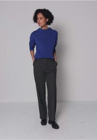 Blauwe gebreide trui met een hoge hals, gecombineerd met donkergrijze, loszittende wollen broek. Model staat in zwarte schoenen tegen een effen achtergrond.