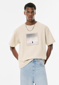 Beige bomulds t-shirt med et firkantet sort-hvidt grafisk print på forsiden, parret med lyseblå jeans. Har løs pasform og rund hals.