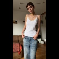 Camisole blanche avec dentelle, associée à un jean bleu clair ample. L'arrière-plan présente un canapé marron et une décoration minimaliste.