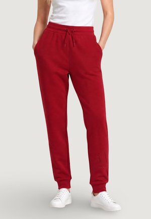 Personne portant un pantalon de survêtement rouge avec taille à cordon et poches, associée à des baskets blanches et un haut blanc sur un fond uni.