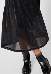 Falda negra con una textura brillante y un dobladillo curvado, combinada con botas de tobillo negras y brillantes. Calcetines gris claro visibles por encima de las botas.