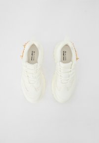 Baskets véganes blanches avec un design texturé, dotées d'une semelle épaisse et d'un laçage à l'avant. Accentuées par un détail en chaîne dorée au niveau du talon.