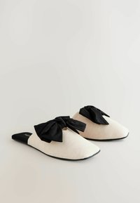 Scarpe slip-on con una parte superiore in morbido tessuto beige, caratterizzate da un fiocco in tessuto nero sulla parte anteriore, punta arrotondata e suola nera piatta.