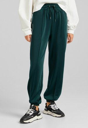Ciemnozielone szerokie spodnie dresowe z elastycznym pasem i nogawkami, zestawione z czarno-białymi masywnymi sneakersami na szarym tle.