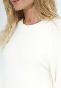 Pull en maille blanche avec un col rond, des poignets côtelés et des coutures avec une texture subtile. Accent sur la douceur et le confort.