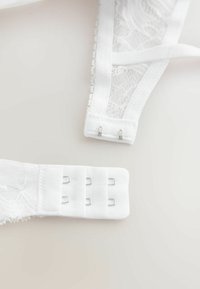 Biały pasek biustonosza z koronki z podwójnym zapięciem na haftki na jednolitej jasnej powierzchni.