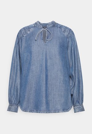 Blouse en denim bleu délavé à manches longues avec col et épaules froncés, encolure en forme de trou de serrure et fermeture à cordon à nouer au niveau du cou.