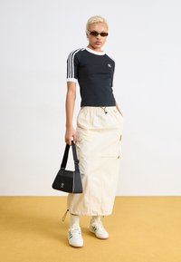 Person med kort blont hår som bär en svart Adidas-tröja, lång beige kjol, vita sneakers, solglasögon och håller en svart Adidas-väska.