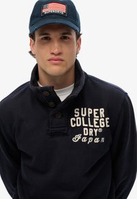 Pullover blu navy con colletto a bottoni, caratterizzato da testo ricamato color crema "SUPER COLLEGE DRY Japan." Cappellino con dettaglio della bandiera americana.