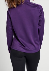 Lila långärmad skjorta i slät tyg med ståkrage och tre vita knappar på axeln, parat med svarta leggings.