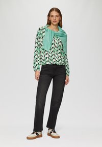 Langemouwen blouse met geometrisch patroon in groen, zwart en wit, gecombineerd met zwarte rechte jeans en crème sneakers met zwarte accenten.