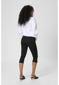 Woman with curly hair wearing a white shirt, black cropped pants, and black heeled slingback shoes, turned slightly to the side on a plain background.