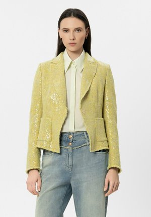 Frau trägt ein hellgelbes, mit Pailletten besetztes Blazer, ein hellgrünes Hemd mit Knöpfen und hoch taillierte hellblaue Jeans vor einem schlichten Hintergrund.