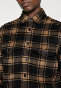 Camicia a quadri nera e marrone, con due taschini sul petto, bottoni a chiusura e colletto. Realizzata in tessuto testurizzato.