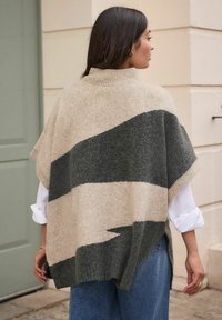 Beige and dark green knitted poncho with a high collar, featuring diagonal stripes, short sleeves, and side slits. Worn over a white shirt.