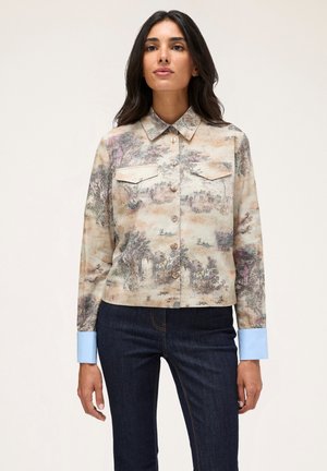 Chemise à manches courtes en tissu beige avec un imprimé paysage, deux poches poitrine et des poignets bleu clair. Boutons à l'avant.