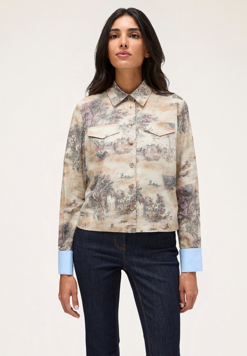 Chemise à manches courtes en tissu beige avec un imprimé paysage, deux poches poitrine et des poignets bleu clair. Boutons à l'avant.