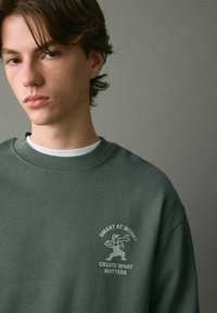 Felpa verde con colletto rotondo realizzata in cotone. Presenta una piccola stampa grafica sul lato sinistro del petto con la scritta "SMART AT WORK® CREA QUELLO CHE CONTA."