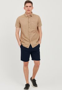 Knytt kortermet beige lin skjorte med brystlomme, parret med marineblå shorts og sorte joggesko. Enkelt design, avslappet passform og oppbrettede ermer.