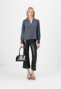 Graue Bluse mit langen Ärmeln und gerafften Manschetten, schwarze Leder-Culotte, schwarze Handtasche und taupefarbene spitze Pumps vor einem neutralen Hintergrund.
