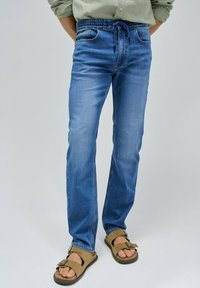 Jeans de mezclilla azul claro con cinturilla elástica y cordón. Cuenta con dos bolsillos frontales y un corte recto y relajado. Llevados con sandalias marrones.