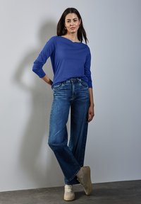 Chemise bleue à manches longues avec un large encolure, associée à un jean taille haute à jambes larges bleu clair. Baskets beiges avec un motif texturé.