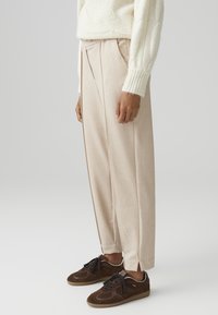 Beige geplooide broeken met een getailleerde pasvorm, gemaakt van lichtgewicht stof. Gecombineerd met donkerbruine suède sneakers met zwarte veters.