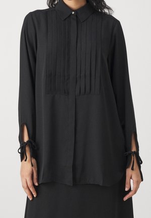 Kobieta ubrana w czarną bluzkę z długim rękawem, z frontowymi zakładkami, wysokim kołnierzem z ozdobnym guzikiem oraz zawiązywanymi mankietami na nadgarstkach.