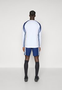 Hvid langærmet skjorte med blå detaljer, parret med blå shorts og sorte knæhøje sokker. Skjorten har en rund halsudskæring og er tætsiddende.