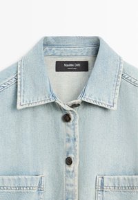 Chemise en denim bleu clair avec un col, deux poches poitrine, fermeture par boutons et une étiquette noire indiquant Massimo Dutti fabriqué en Turquie.