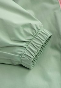 Tessuto verde chiaro con texture liscia, caratterizzato da polsini elasticizzati arricciati. Il materiale ha una leggera lucentezza e un fine motivo a griglia.