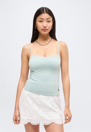 Young woman with long dark hair wearing a light blue spaghetti strap top and white eyelet lace skirt, standing against a plain background.