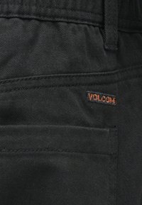 Svarte stoffbukser med en subtil tekstur, en baklomme, og en liten oransje "Volcom"-logo brodert på et lag.