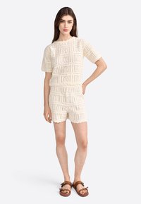 Conjunto de dos piezas de color crema de punto, que incluye una camiseta de manga corta y pantalones cortos con intrincados patrones de calados y bordes ondulados.