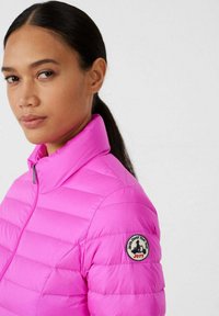 Giacca imbottita rosa con collo alto, dotata di chiusura con zip e toppa rotonda con logo sul braccio sinistro. Tessuto liscio e lucido, design aderente.