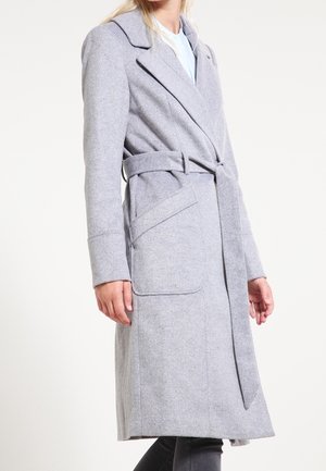 Kvinde iført en lang, lysegrå uldfrakke med bælte, brede revers og forlommer med klap, kombineret med mørke bukser.