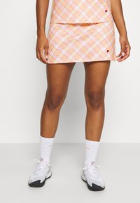 Falda atlética de cuadros en naranja y rosa pastel, hecha de un tejido ligero. Presenta un logo en la cadera izquierda y se combina con zapatillas blancas y calcetines.