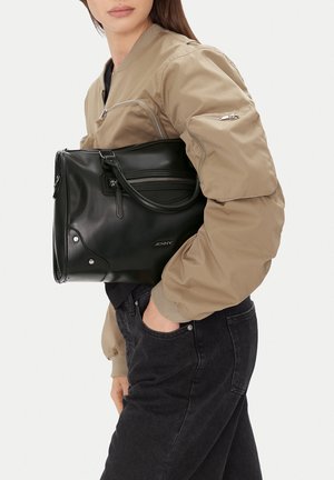 Donna che indossa una giacca bomber beige e jeans neri, che tiene una borsa a tracolla in pelle nera con cerniera e logo "JENNY".