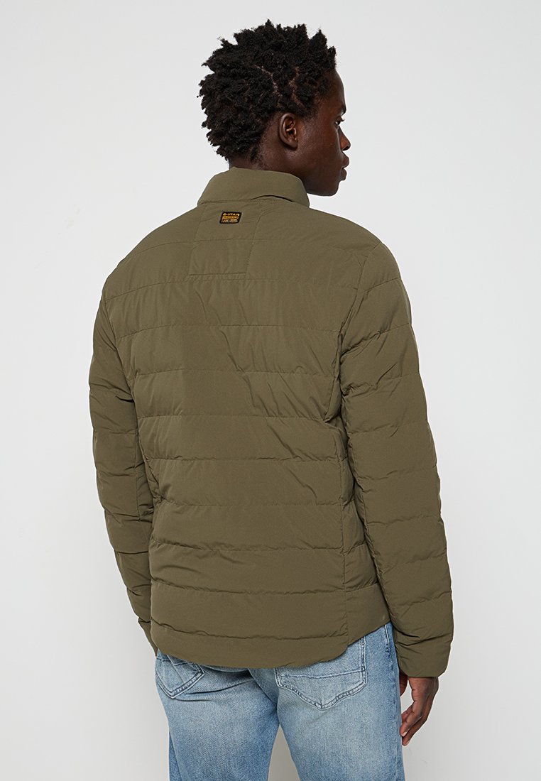 Hombre de espaldas con una chaqueta acolchada verde olivo y jeans de color azul claro frente a un fondo sencillo.