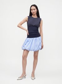 Femme debout portant un haut sans manches bleu marine avec une mini-jupe bouffante bleu clair et des chaussures blanches à talons bas et à bride arrière, sur un fond uni.