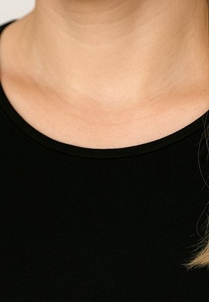 Nahaufnahme des Halses und oberen Brustbereichs einer Person, die ein schwarzes Rundhalsshirt trägt, mit einem kleinen Strang hellem Haar sichtbar auf der rechten Seite.