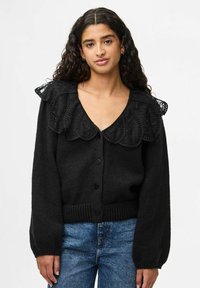 Melnā adīta kardigāna ar dziļu V-veida kakla izgriezumu, dekortīvu mežģīņu frills apkakli un pogu aizdari. Iezīmes ribotas piedurknes un apakšmalu.