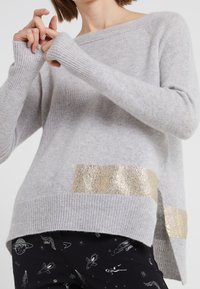 Harmaa neulepusero, jossa on raidoitetut hihansuut, sisältäen kullanvärisen metallisen yksityiskohdan alhaalla ja halkiotyylin helman.