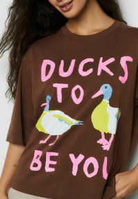 Tricou maro din bumbac cu design grafic ce reprezintă două rațe colorate și textul „DUCKS TO BE YOU” în roz. Mâneci scurte și croială lejeră.