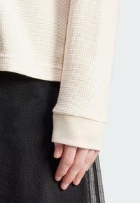 Top de manga larga texturizado color beige claro con cuello y puños acanalados, combinado con una falda de mezclilla negra, mostrando una mano apoyada en la tela.