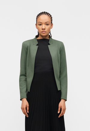 Vrouw met gevlochten haar draagt een groene, op maat gemaakte blazer over een zwart topje en een geplooide zwarte rok, staand tegen een effen witte achtergrond.