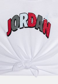 T-shirt bianco con orlo annodato. Presenta una grafica "JORDAN" in bold multicolore nei toni rosso, grigio e blu, con una texture marmorizzata.