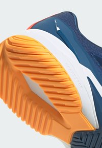 Urheilujalkineen pohja, jossa on teksturoitua oranssia kumia valkoisilla yksityiskohdilla, mikä tarjoaa pidon. Tummansininen yläosa, jossa on kiiltävä sininen suunnitteluelementti.
