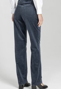 Osoba w ciemnoniebieskich sztruksowych spodniach, białych koszulowych rękawach i czarnych butach na obcasie, stojąca na tle jasnoszarym.